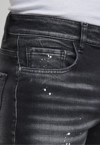 Gros plan sur un jean en denim noir avec des éclaboussures de peinture blanche et une texture usée, porté avec un t-shirt blanc côtelé rentré à l’intérieur.