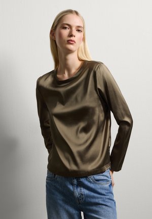 Camicia lunga in satin verde oliva con scollo rotondo, texture liscia e vestibilità rilassata, abbinata a jeans di denim azzurri chiari.