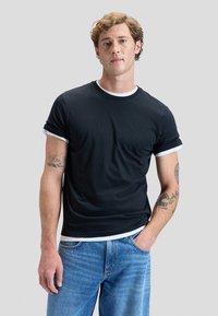 Ung mand med krøllet hår iført en sort T-shirt med hvide kanter og blå jeans, stående med den ene hånd i lommen mod en ensfarvet baggrund.