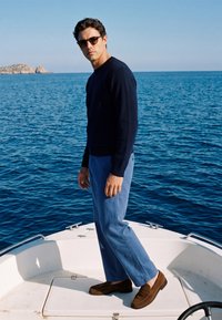 Homme portant des lunettes de soleil, un pull noir, un pantalon bleu et des mocassins marron, debout sur le pont d'un bateau avec une mer bleue calme et une côte rocheuse lointaine derrière.