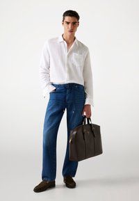 Brown leather tote bag with gold hardware, white linen shirt, blue denim pants, and dark loafers with fringe detailing.
