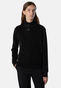 Chaqueta negra con cremallera en tejido suave y cepillado. Presenta un cuello alto, bolsillos laterales y un sutil acento de logo en el puño. Textura suave.