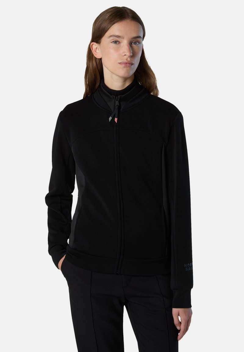 Chaqueta negra con cremallera en tejido suave y cepillado. Presenta un cuello alto, bolsillos laterales y un sutil acento de logo en el puño. Textura suave.