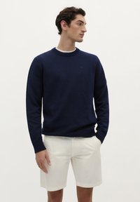 Marineblå strikket sweater med rund ribbed halsudskæring, lange ærmer og ribbet kant, parret med hvide shorts. Glat tekstur, afslappet design.