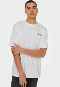 Witte katoenen t-shirt met korte mouwen, met "London Fields" geborduurd in groene en rode tekst op de linker borst.