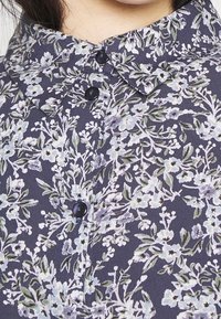Camicia a bottoni floreale viola con fiori blu e bianchi, foglie verdi e un classico design del colletto con bottoni scuri.