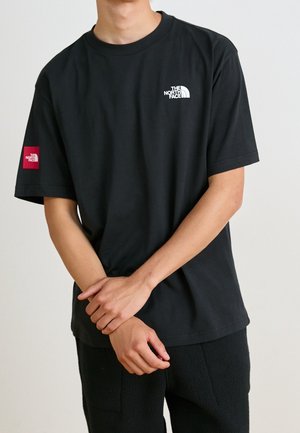 Personne portant un t-shirt noir The North Face avec logo blanc sur la poitrine et logo patch rouge sur la manche gauche, les mains jointes devant.