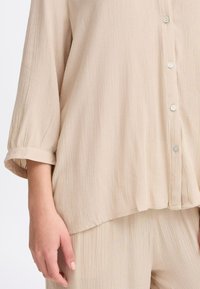 Šviesiai rudas mygtukų mygtukais marškinis su tekstūruotu, susiraukšlėjusiu audiniu. Sukurta su ilgomis rankovėmis ir sukurtomis rankogaliais, taip pat baltais mygtukais palei apykaklę.