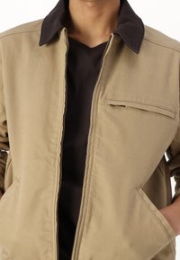 Chaqueta beige con un cuello oscuro y cremallera frontal. Cuenta con un bolsillo en el pecho y bolsillos laterales. La tela tiene una textura suave y estructurada.