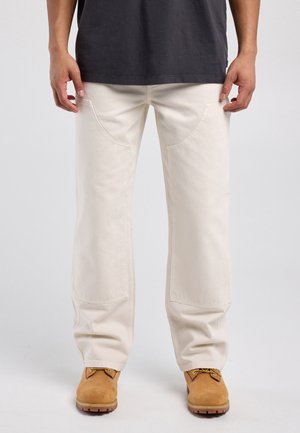 Persoon die off-white rechte broek met grote voorzakken, tan werkschoenen en een donkergrijze shirt draagt, staand tegen een lichte achtergrond.