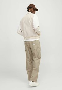 Chaqueta bomber beige con mangas blancas, pantalones cargo beige y zapatillas blancas. La gorra marrón agrega contraste. Diseño simple y funcional.