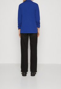 Modrá dlouhá bunda s ohrnutými rukávy v kombinaci s černými kalhotami se vzorem malých teček a černou obuví, z pohledu zezadu.