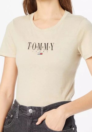 Beige T-shirt met korte mouwen met de tekst en het logo "TOMMY 1985 JEANS", gecombineerd met zwarte high-waisted jeans, hand in de zak.