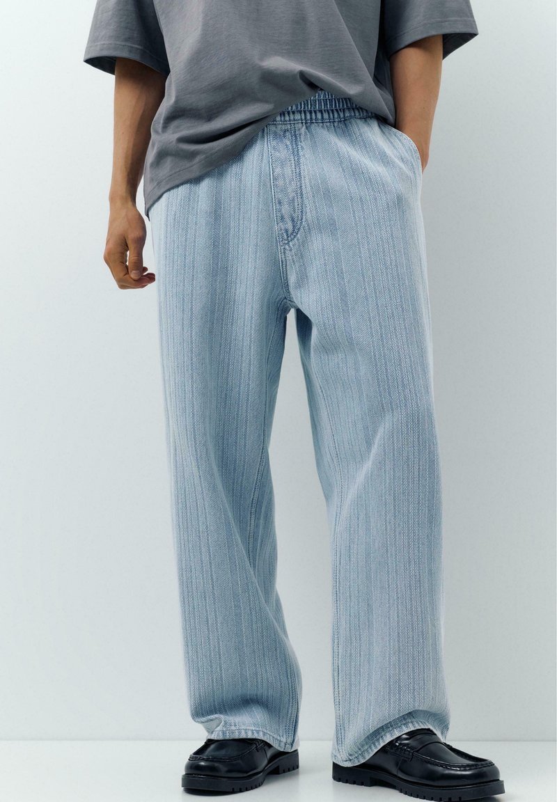 Osoba ubrana w luźne, jasnoniebieskie spodnie w paski z elastycznym pasem, szarą koszulkę i czarne skórzane buty na jednolitym tle.