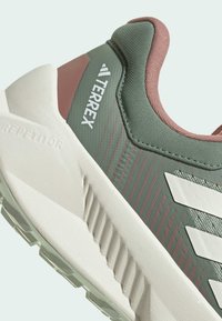 Scarpa sportiva Terrex con una parte superiore verde testurizzata e dettagli in rosa, strisce bianche e una suola bianca stampata con dettagli antiscivolo.