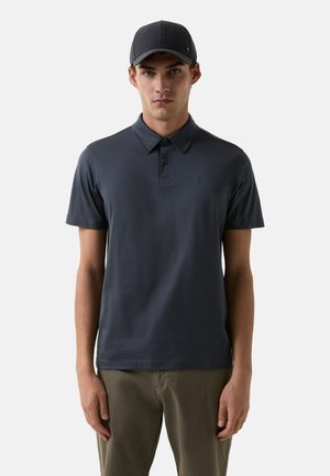 Marineblå polo-shirt med krave, korte ærmer og en diskret logo-detalje. Stylet med lyse bukser. Glat bomuldsstruktur.
