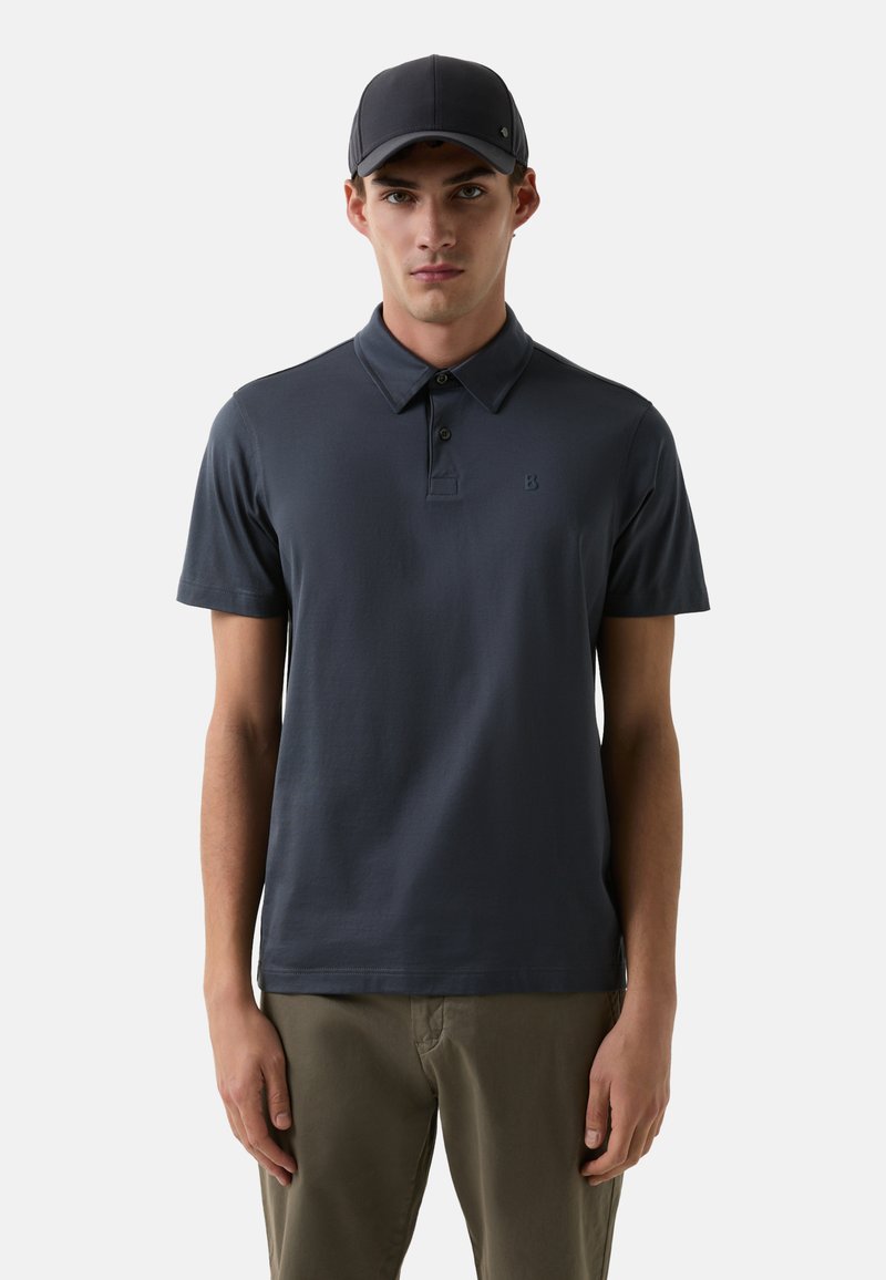 Marineblå polo-shirt med krave, korte ærmer og en diskret logo-detalje. Stylet med lyse bukser. Glat bomuldsstruktur.