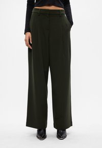 Γυναίκα με ψηλόμεσες, φαρδιές σκούρες πράσινες παντελόνες με πτυχώσεις μπροστά, μαύρη μακρυμάνικη μπλούζα και μαύρες μπότες με κοφτές μύτες.