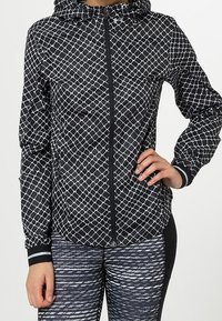 Woman wearing black and white geometric patterned zip-up jacket and black and white patterned leggings, with one hand on hip.