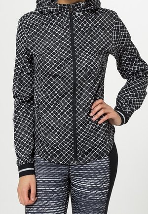 Kobieta ubrana w czarną i białą kurtkę z geometrycznym wzorem na zamek oraz legginsy z czarno-białym wzorem, z jedną ręką na biodrze.