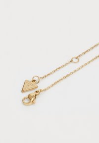 Goldene Halskette mit Kettendesign, ausgestattet mit einem dreieckigen Anhänger, der mit "GUESS" geprägt ist, und einem Karabinerverschluss zur Sicherung.