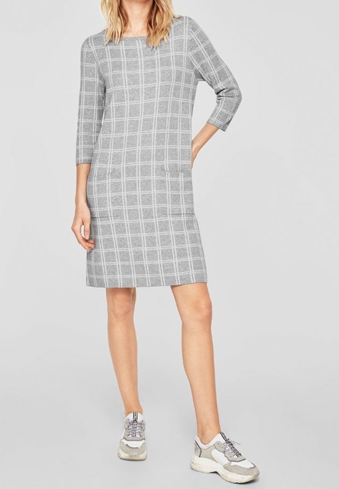 Femme portant une robe grise à carreaux longueur genou avec des manches trois-quarts et des baskets grises et blanches décontractées, debout avec une main sur la hanche.