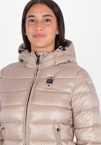 Giubbotto imbottito beige con cappuccio, caratterizzato da un design a quadrati orizzontali, chiusura con zip anteriore e toppa con logo sulla manica sinistra.