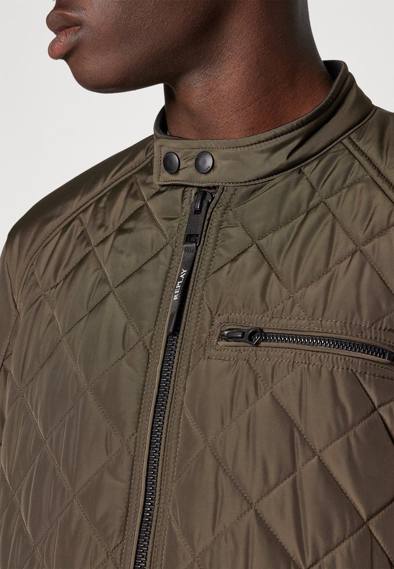 Nahaufnahme einer Person, die eine olivgrüne Steppjacke mit Reißverschlussfront, Brustreißverschlusstasche und Druckknopfkragen trägt.