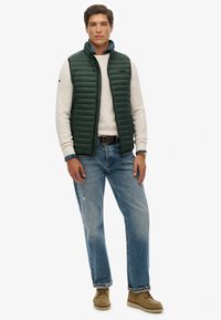 Groene gewatteerde bodywarmer over een witte gebreide trui, gecombineerd met lichtblauwe jeans en bruine laarzen. Voorzien van een geruite boord van het overhemd en voren zakken aan de voorkant.