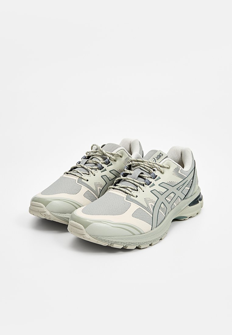 Une paire de chaussures de course ASICS grises et beiges avec des lacets, un dessus en tissu en maille et des semelles en caoutchouc sur un fond blanc.