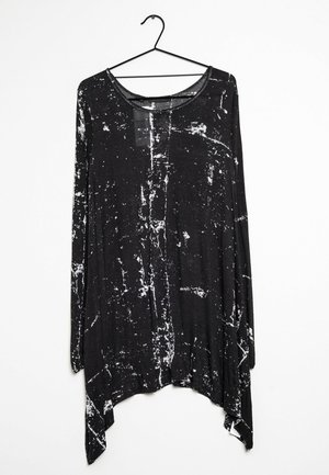 Chemise noire à manches longues avec ourlet asymétrique et motif éclaboussé abstrait blanc, suspendue sur un cintre en fil noir contre un mur blanc.