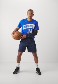 Modrá športová tričko s nápisom "MILWAUKEE 34" vytlačeným bielou farbou, kombinované s námořníckymi šortkami. Čierna rukávová manžeta, drží hnedý basketbal. Čierne tenisky.