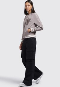 Veste bomber gris clair avec broderie, poignets rayés et poches zippées, associée à un pantalon cargo noir et des baskets blanches.
