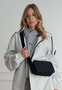 Černá obdélníková crossbody taška s hladkým povrchem a embosovaným logem, nošená přes šedou mikinu a bílou nepromokavou bundu.