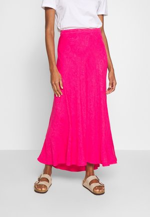 Femme portant une jupe longue maxi rose vif à motifs, un t-shirt blanc et des sandales beige à double brides, debout devant un fond clair uni.
