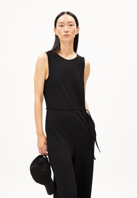 Schwarzer ärmelloser Jumpsuit mit taillierter Silhouette, gerafftem Taillenzug und rundem Halsausschnitt. Das Model hält eine schwarze Baseballmütze.