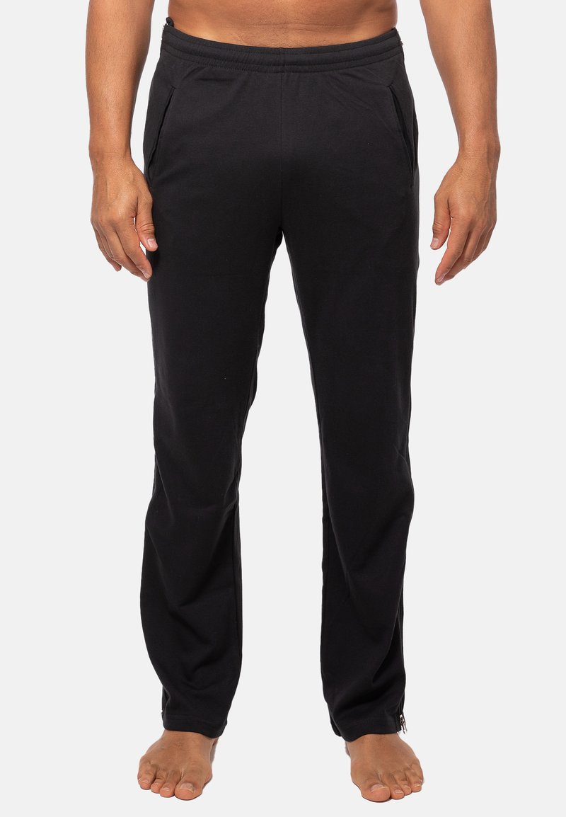 Homme portant un pantalon droit noir avec des poches latérales, debout pieds nus contre un fond blanc uni.