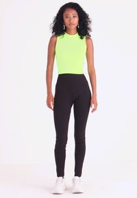 Top sem mangas verde néon combinado com leggings pretas de cintura alta, ambos feitos de tecido elástico, usados com ténis brancos.