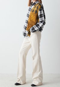Chemise en flanelle à carreaux noir et blanc avec une poche tricotée jaune ornée de franges. Pantalon évasé de couleur crème et chaussures noires à talons.