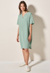 Hellgrünes Leinenkleid mit V-Ausschnitt und kurzen Ärmeln, lockere Passform. Kombiniert mit weißen Sneakers. Einfaches und modernes Design.