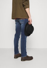 Tummanvihreä takki punaisella ruudullisella paidalla, siniset kapeat farkut ja ruskeat nahkakengät. Musta huopahattu pidetään oikeassa kädessä.