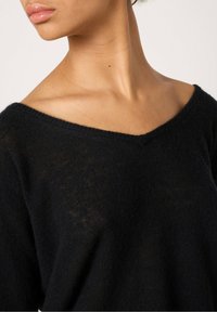 Pull en maille noir avec un décolleté en V, texture douce et coupe décontractée, laissant entrevoir une touche de peau sur les épaules.