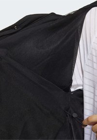 Černá síťovaná vesta se zipem vpředu a stahovací šňůrkou, s texturovaným povrchem a lehkým materiálem, vrstvená přes bílé pruhované tričko.