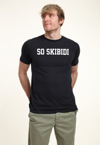 Černé tričko s krátkým rukávem vyrobené z bavlněné směsi s bílým nápisem "SO SKIBIDI" tučným písmem přes hrudník. Jednoduchý design, přiléhavý střih.
