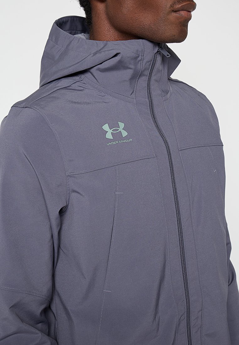 Veste légère grise avec capuche, dotée d'une fermeture éclair à l'avant, de coutures subtiles et du logo Under Armour en vert clair sur le haut de la poitrine gauche.