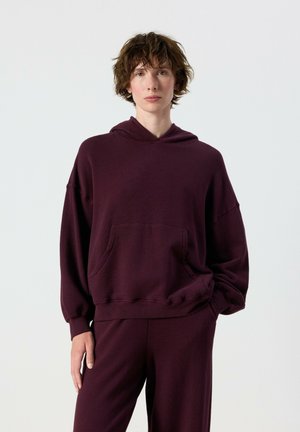 Person with short curly hair wearing a loose dark purple hoodie and matching pants, standing against a plain light background.
