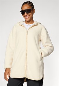 Lächelnde Person in einer cremefarbenen Fleecejacke und großen Sonnenbrillen, die Hände in den Taschen, vor einem hellgrauen Hintergrund.