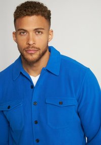 Blå fleece skjorte med spidse kraver, sorte knapper og to brystlommer med klapper, der giver en blød tekstur og et afslappet design.