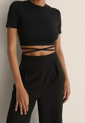 Vrouw in een zwarte cropped top met korte mouwen en kruisbanden rond de taille, die hoge zwarte broek draagt en tegen een lichte muur staat.