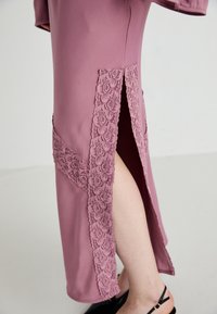 Vestito lungo e aderente in rosa pastello con dettagli in pizzo floreale e spacchi laterali. Tessuto morbido, maniche corte e scollo discreto.